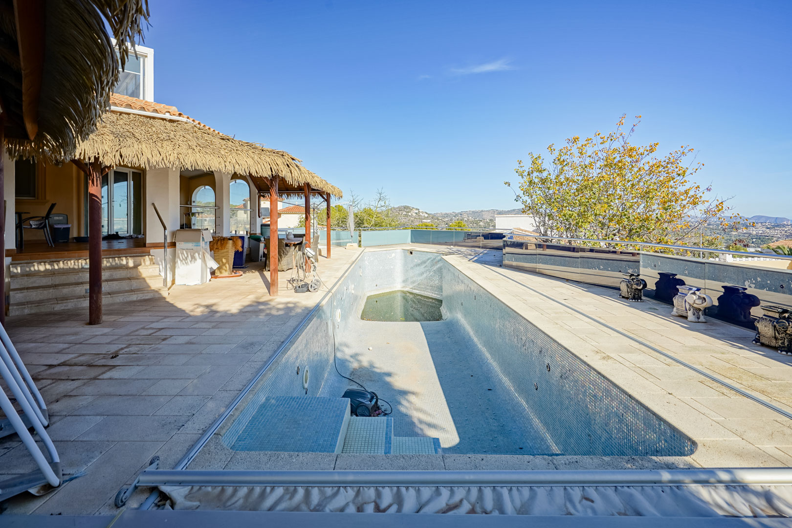Außenpoolbereich mit Stein-Solarium, rustikaler Reetdach-Pergola und Glasgeländern mit weitem Blick auf die umliegende Berglandschaft.