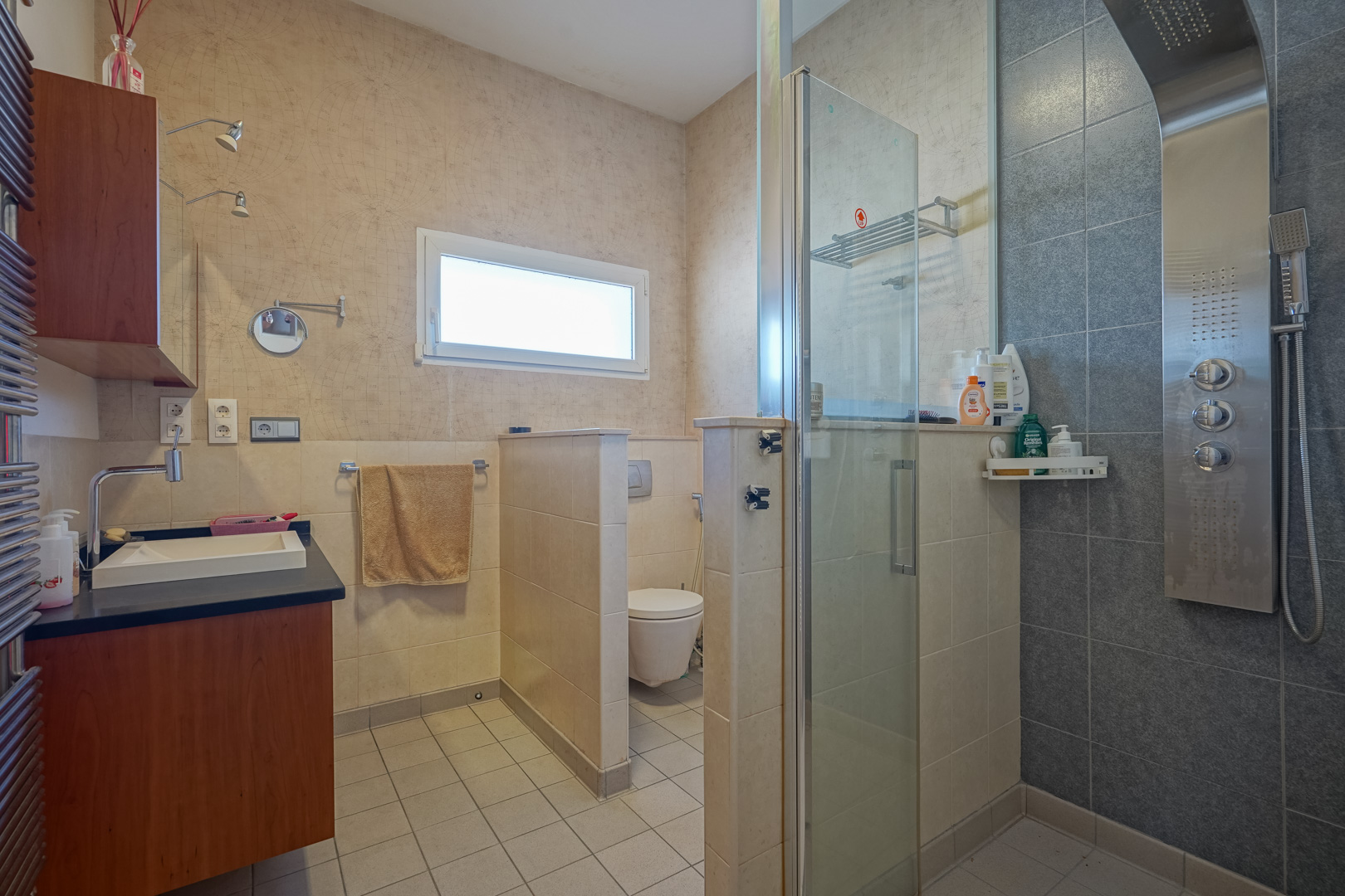 Badezimmer mit Hydromassage-Duschpaneel, Glastrennwand und separatem WC. Ausgestattet mit Holzwaschtisch, dunkler Arbeitsplatte und Tapete mit Weltkartenmuster.