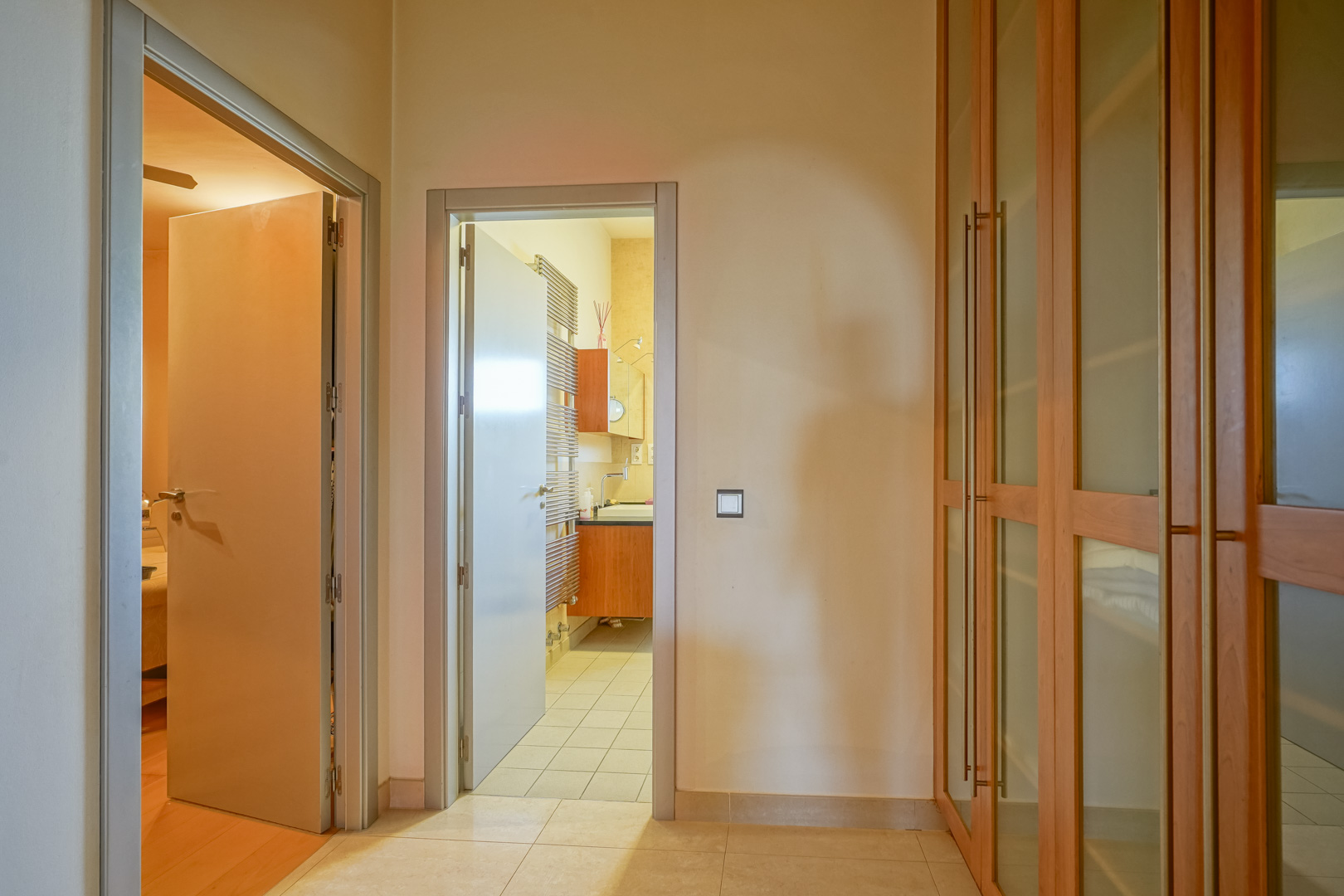 Flurbereich mit Einbauschränken aus Holz und Glas, der ein Schlafzimmer mit Edelholzparkett mit einem En-Suite-Badezimmer inklusive Handtuchheizkörper verbindet.