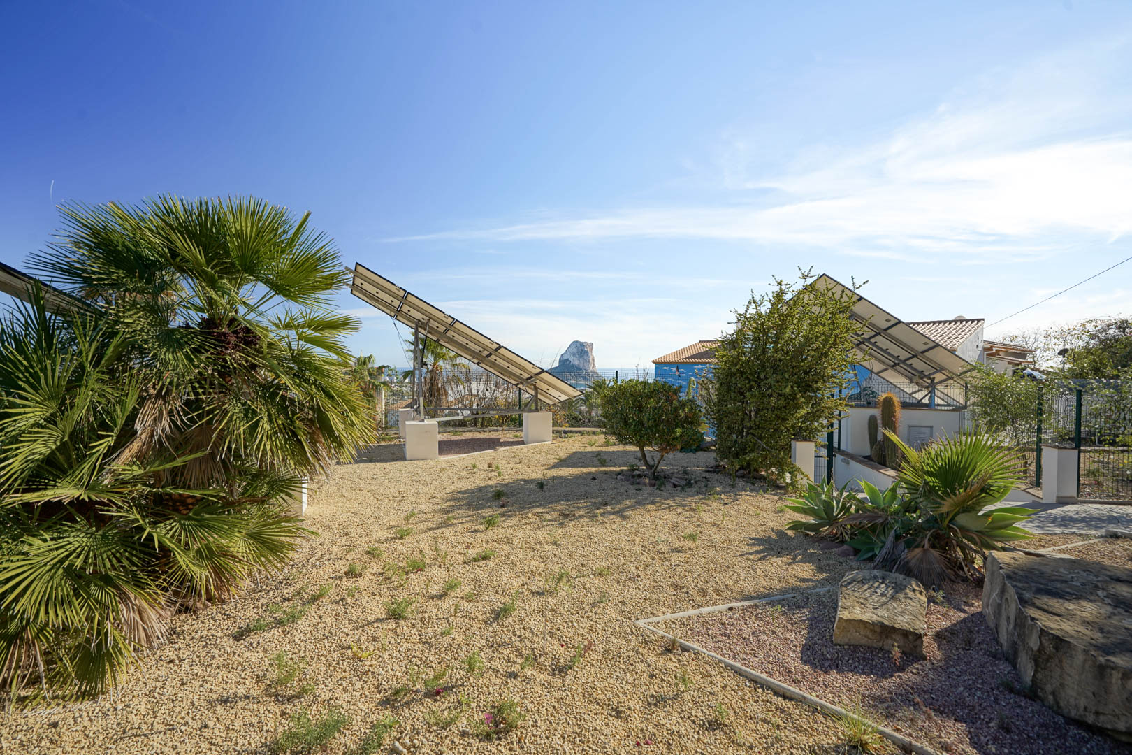 Fortschrittliche Photovoltaikanlage mit motorisierten Solartürmen in einem Kiesgarten, mit Blick auf den Peñón de Ifach und mediterrane Flora.