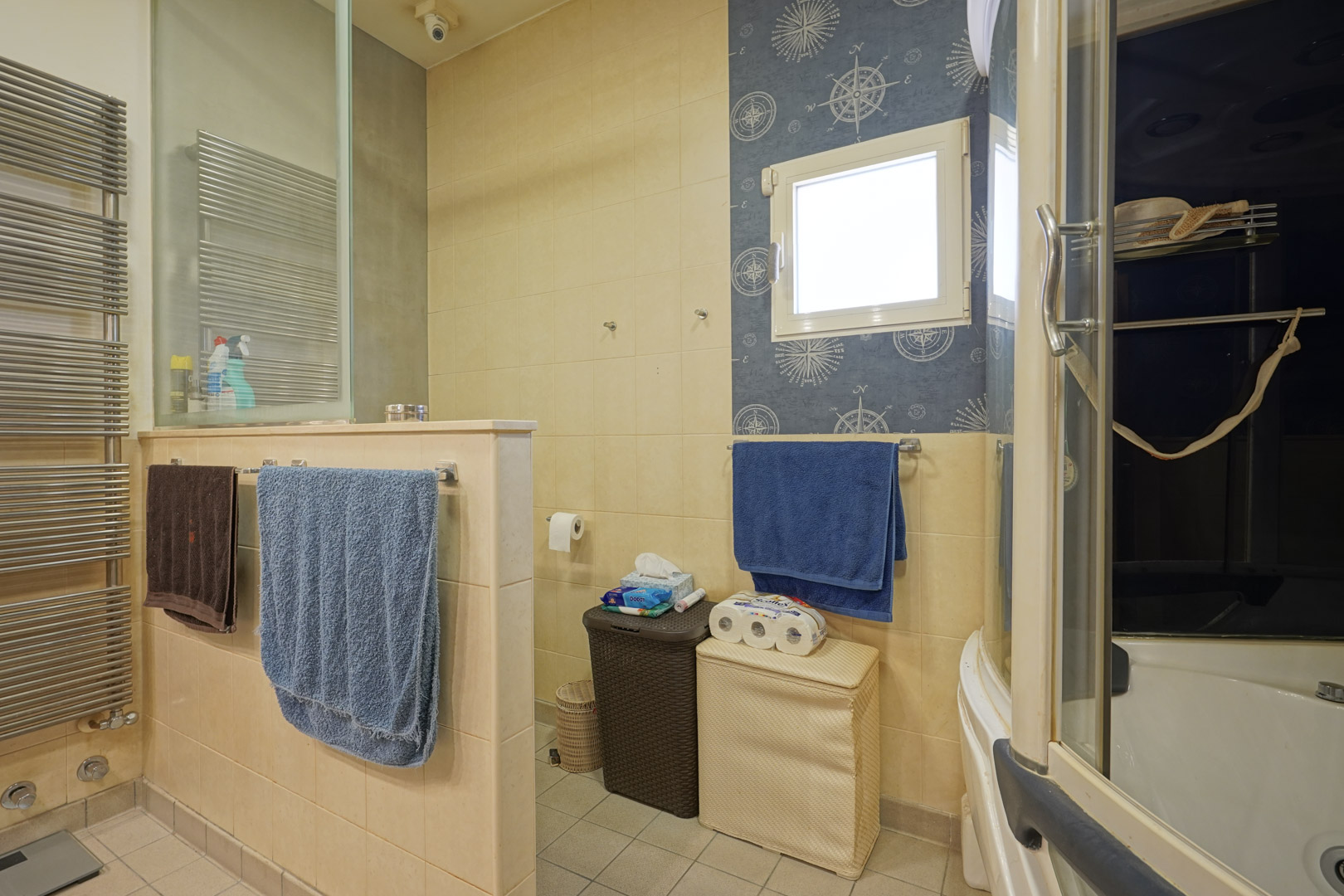 Funktionales Badezimmer mit cremefarbenen Fliesen, Handtuchheizkörper aus Edelstahl und separatem WC-Bereich. Ein nautisches Akzentdekor rahmt das Fenster ein.