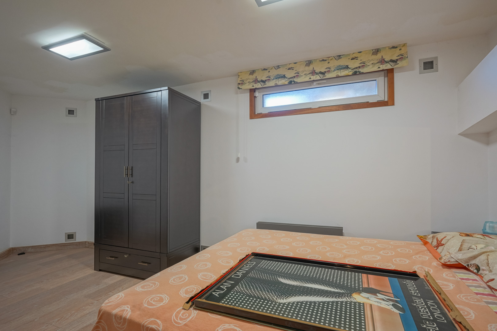 Funktionales Schlafzimmer mit einem dunklen Kleiderschrank, hellem Laminatboden und einem horizontalen Fenster unter quadratischen Deckeneinbauleuchten.