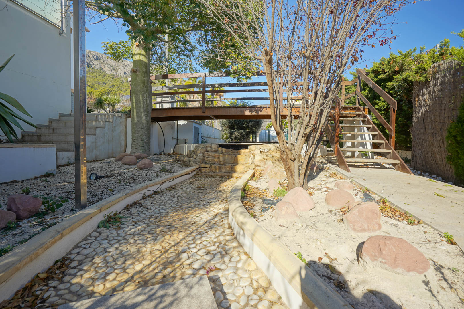 Gartenbereich mit einer Holzbrücke über einem dekorativen Steinwasserfall, einem gepflasterten Kieselweg und Treppen vor der Kulisse der Oltamar-Berge.