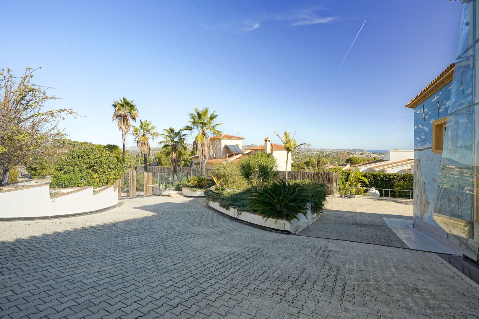 Gepflasterte Einfahrt mit Verbundsteinen, einer bepflanzten Garteninsel, Palmen und einem Wandgemälde, mit weitem Blick auf die Küstenlandschaft von Calpe.