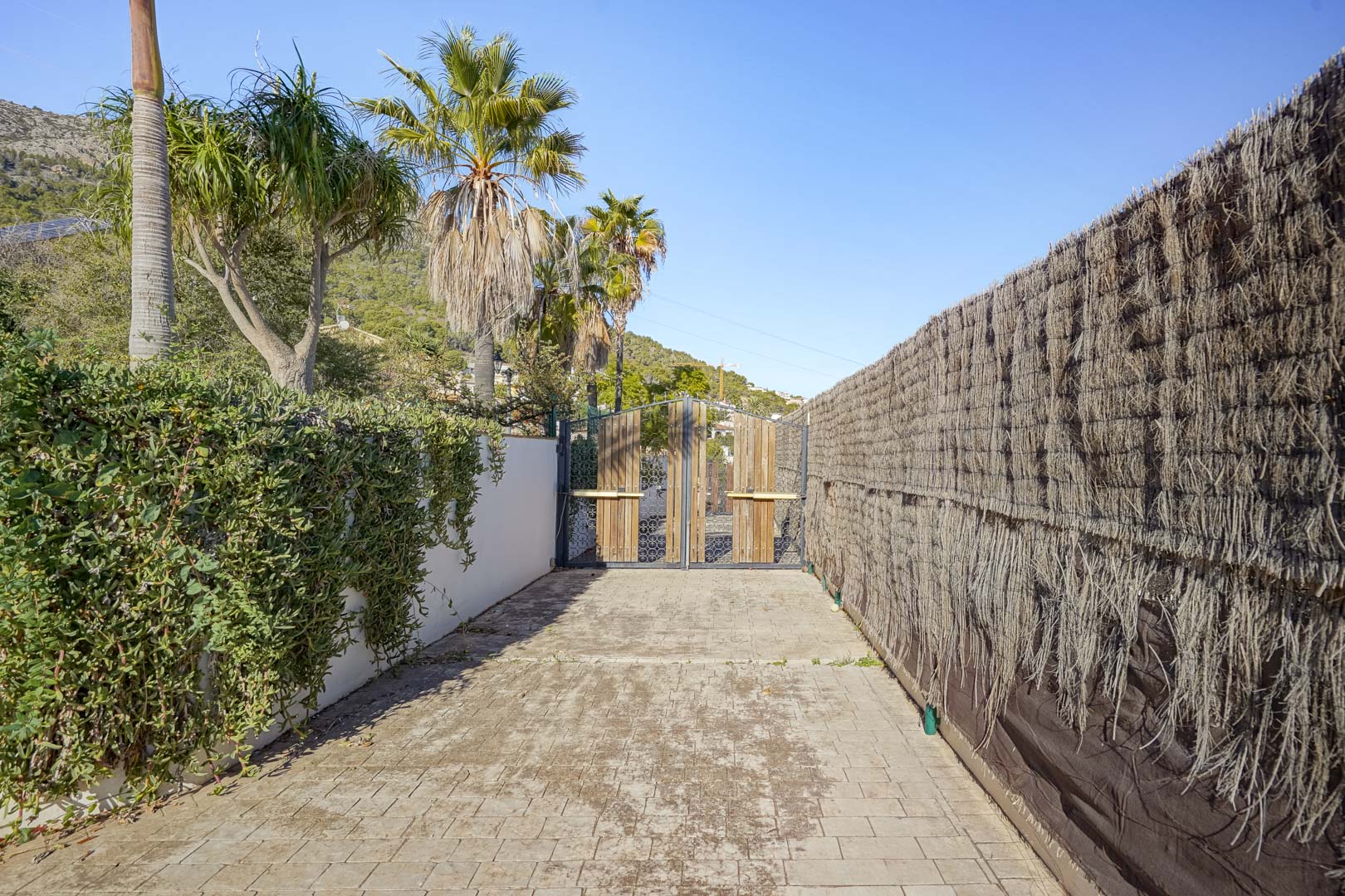 Grundstückszufahrt mit gepflastertem Weg und Holztor, umgeben von mediterraner Bepflanzung, Palmen und einem Sichtschutzzaun aus Heidekraut unter blauem Himmel.