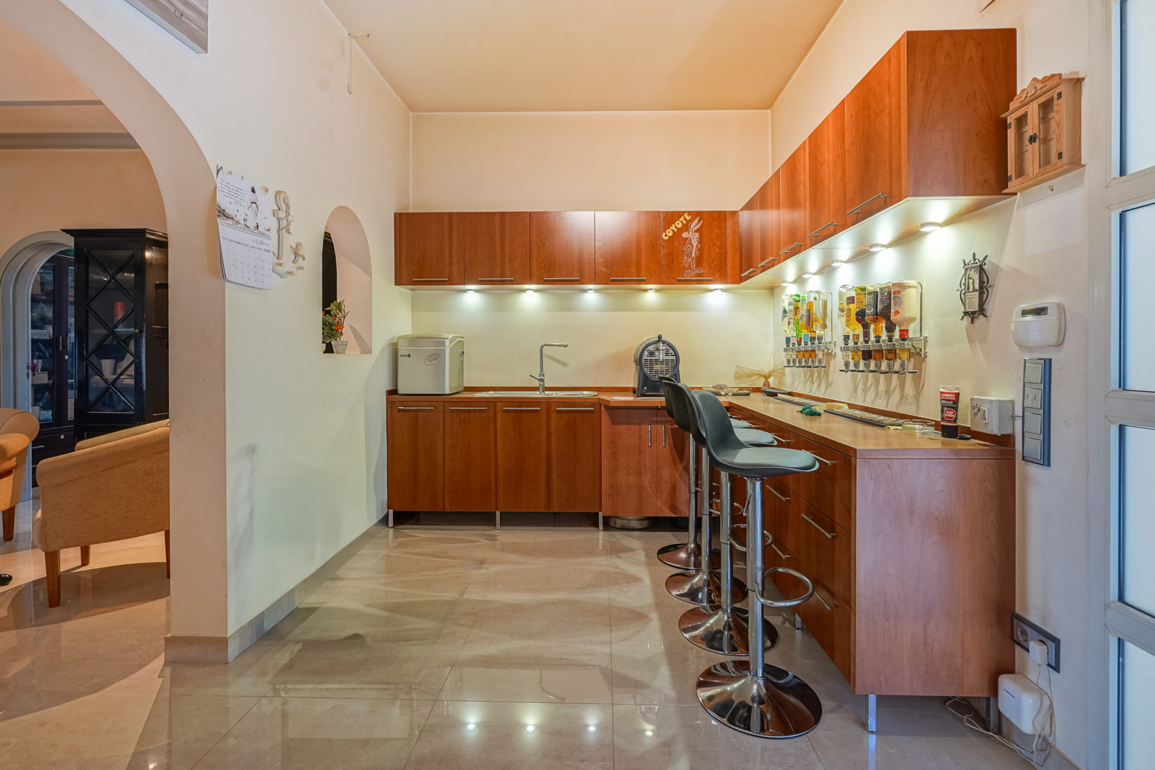 Integrierte Bar mit Holzschränken, Spülbecken und Wand-Getränkespendern. Mit modernen Barhockern, LED-Unterbaubeleuchtung und poliertem Fliesenboden ausgestattet.