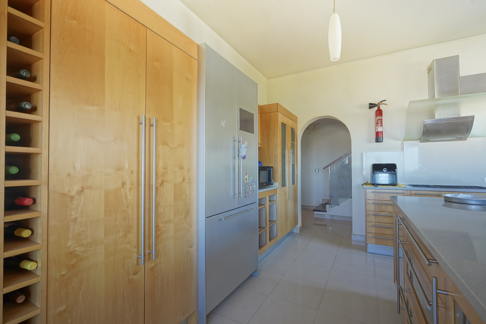 Küche mit maßgefertigten Holzschränken, integriertem Weinregal und Edelstahlelektrogeräten. Ein Rundbogen führt zum Treppenhaus mit modernem Glasgeländer.