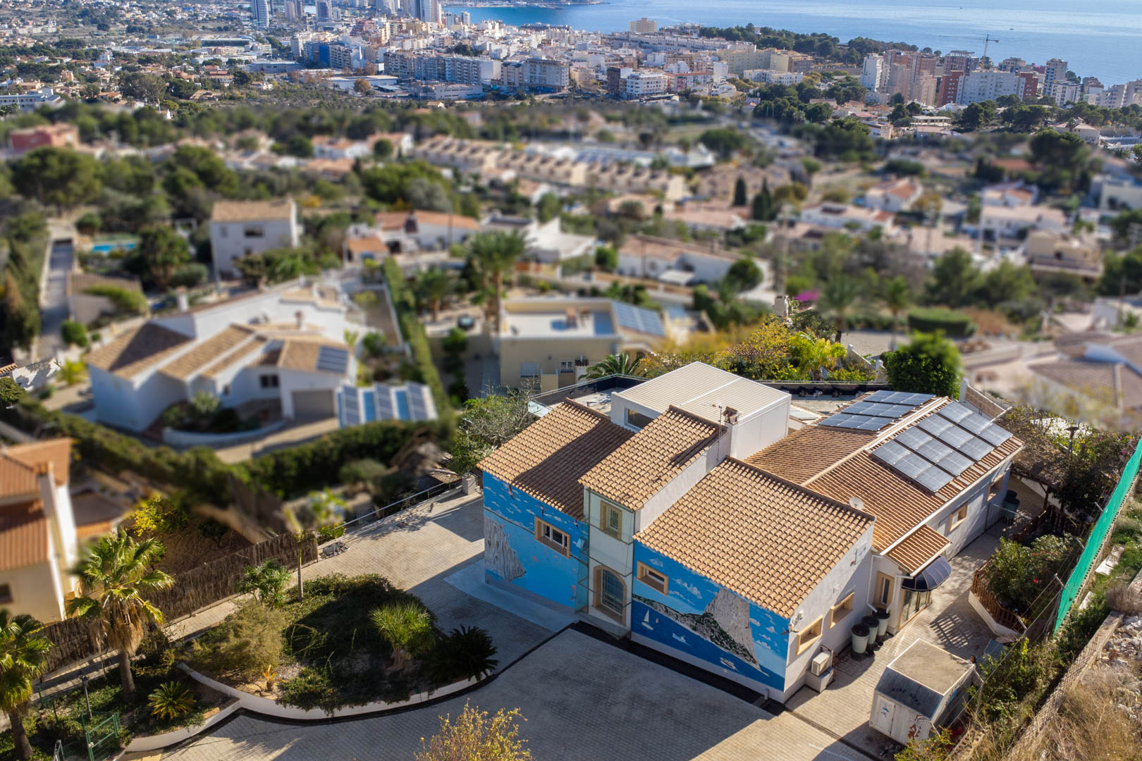 Luftaufnahme der Villa mit kunstvollen Wandmalereien, Ziegeldach und Solaranlage, gelegen in einem Wohngebiet mit Blick auf die Küste und Skyline von Calpe.