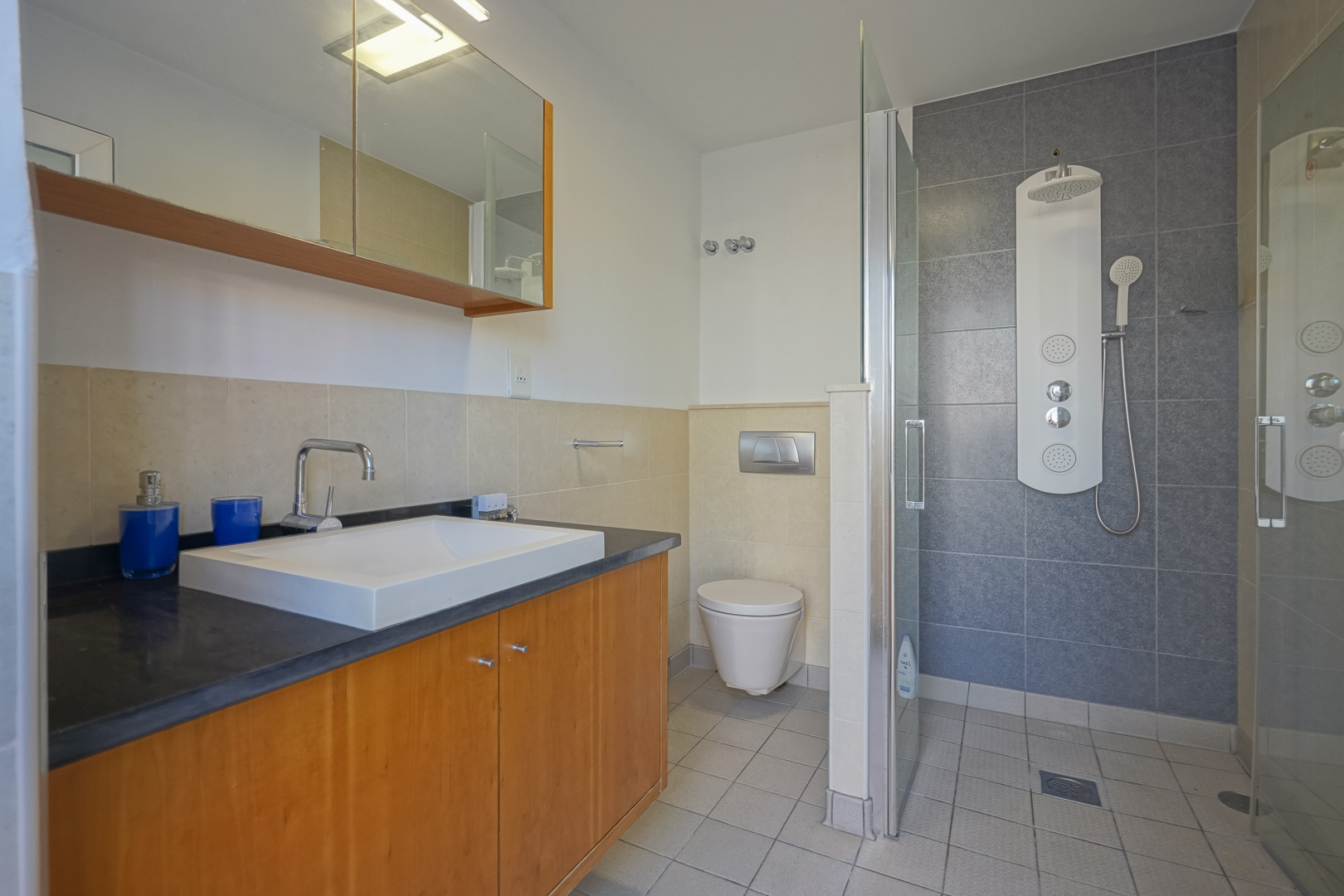 Modernes Badezimmer mit Holzwaschtisch, dunkler Arbeitsplatte und begehbarer Dusche. Ausgestattet mit einer Hydromassage-Säule, Glastrennwand und neutraler Fliesengestaltung.