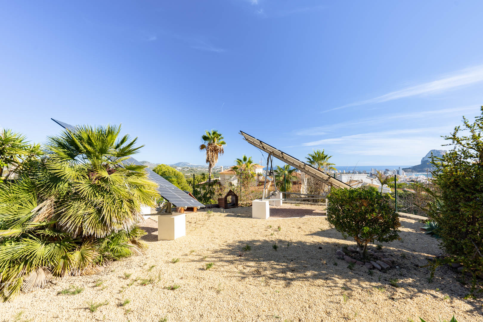 Motorisierte Solaranlagen auf Betonsockeln in einem Kiesgarten mit Panoramablick auf den Peñón de Ifach und die Skyline von Calpe.