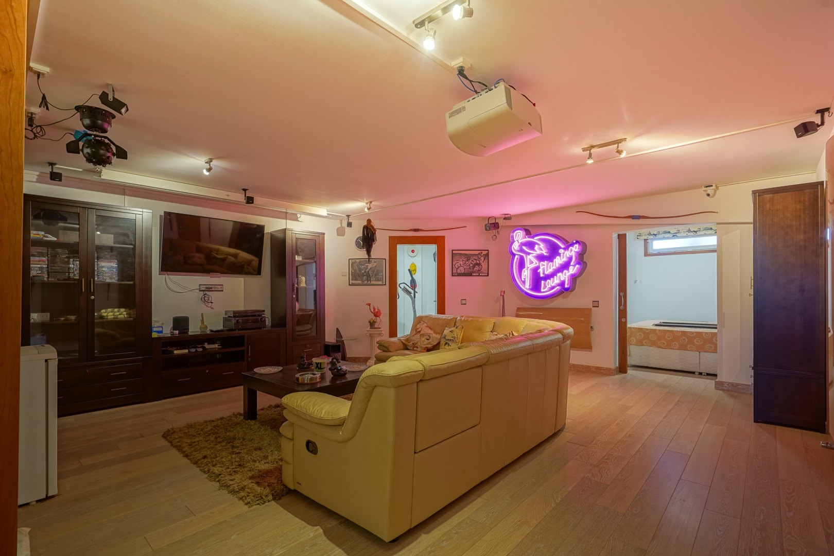 Multimediarraum mit Holzboden, Deckenprojektor und Ledersofa. Der Bereich umfasst dunkle Holzschränke, Schienenbeleuchtung und einen dekorativen Neon-Schriftzug an der Wand.