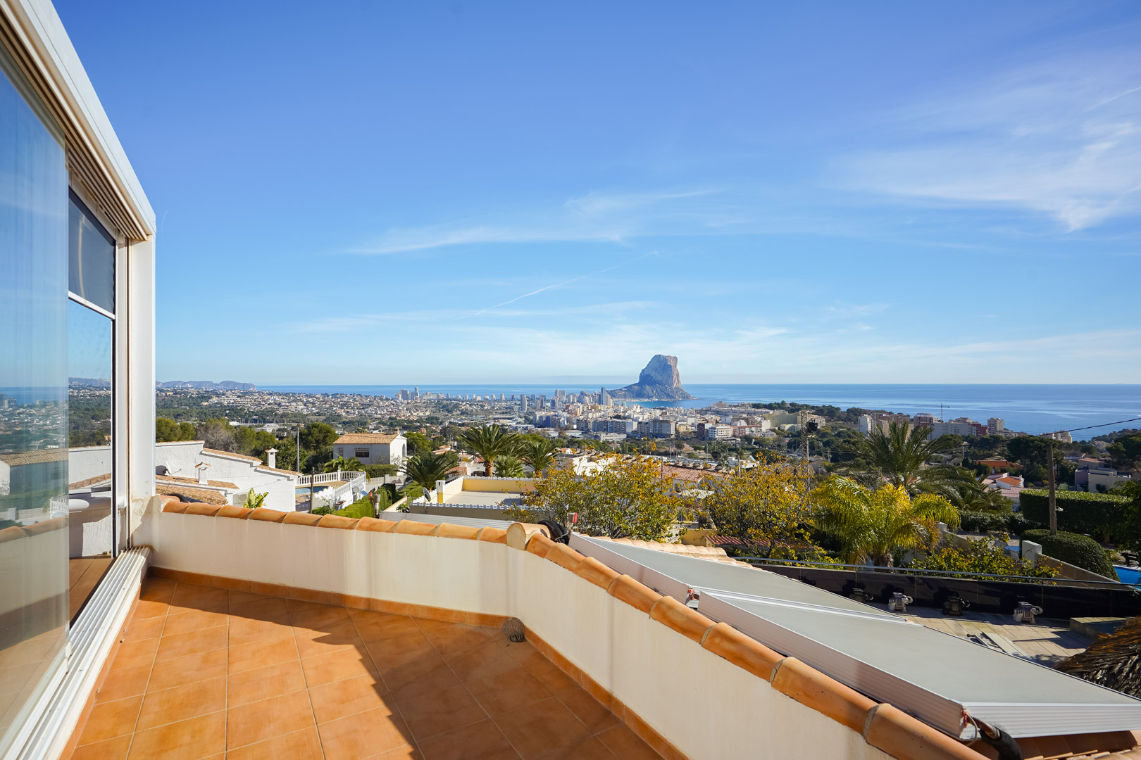 Obere Terrasse mit Terrakotta-Fliesen und Panoramablick auf den Peñón de Ifach und die Skyline von Calpe. Photovoltaikanlagen sind auf dem darunter liegenden Dach sichtbar.