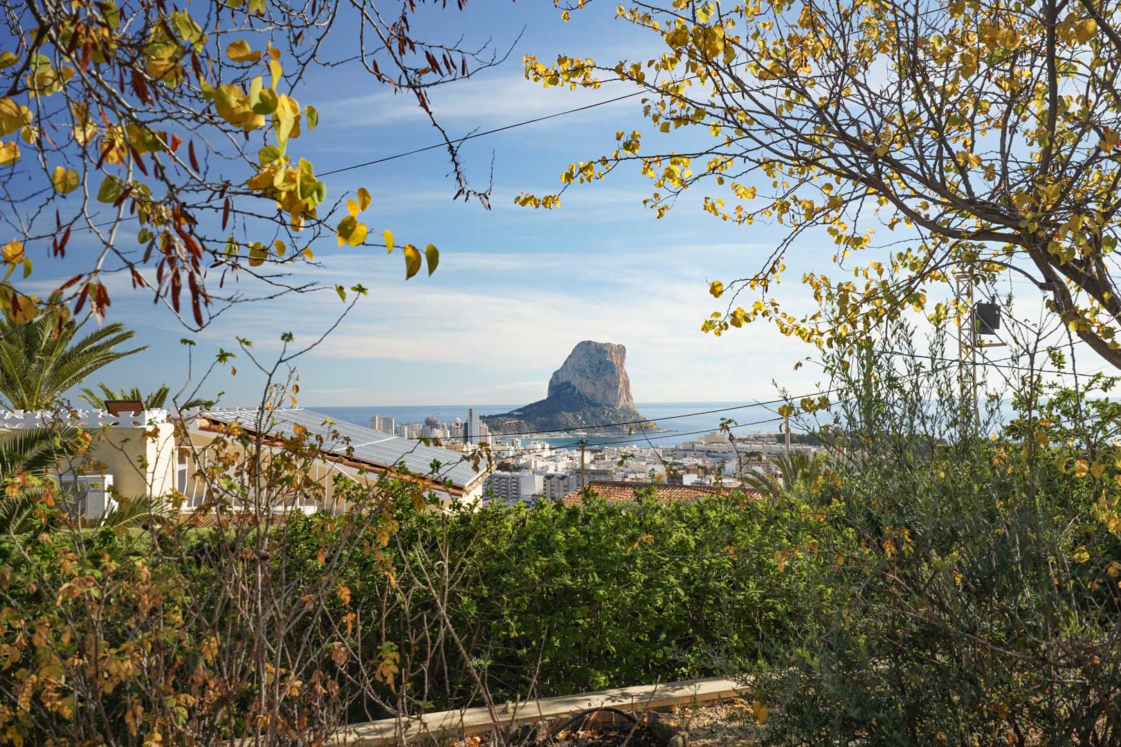 Panoramablick auf den Peñón de Ifach und die Skyline von Calpe, mit Fokus auf die Photovoltaikanlage auf dem Dach inmitten der mediterranen Gartenlandschaft.