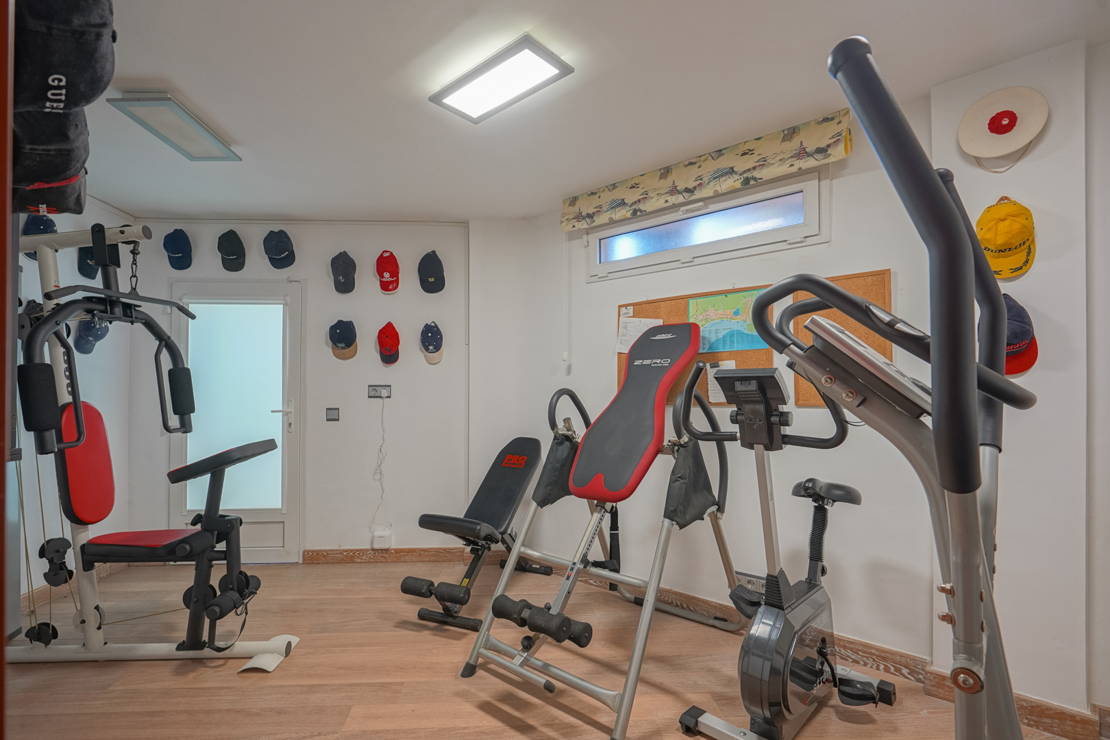 Privater Fitnessraum mit Holzboden, ausgestattet mit Kraftstation, Heimtrainer und Inversionsbank unter flachen LED-Deckenleuchten.