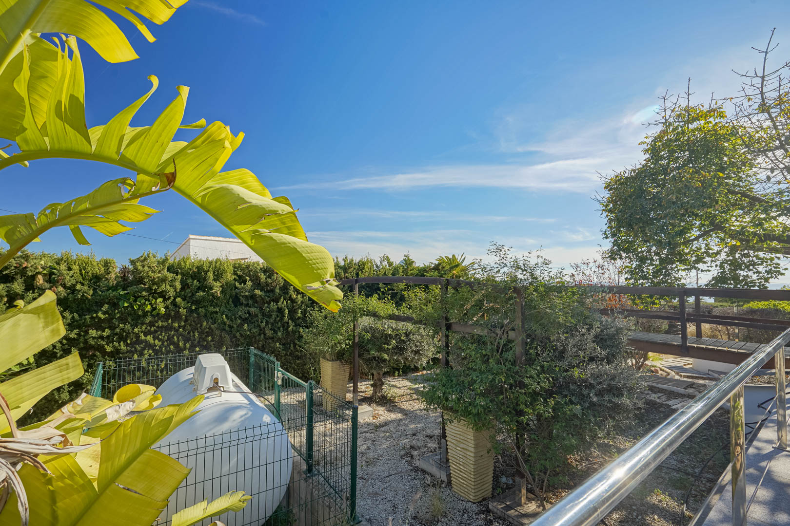 Seitlicher Gartenbereich mit eingezäuntem Gastank, einem Holzsteg und tropischer Bepflanzung unter blauem Himmel.