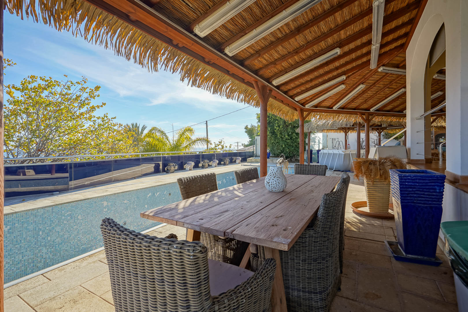 Überdachte Terrasse mit rustikalem Holzesstisch und Korbstühlen unter einer Reetdach-Pergola, direkt am Pool mit Blick ins Grüne gelegen.