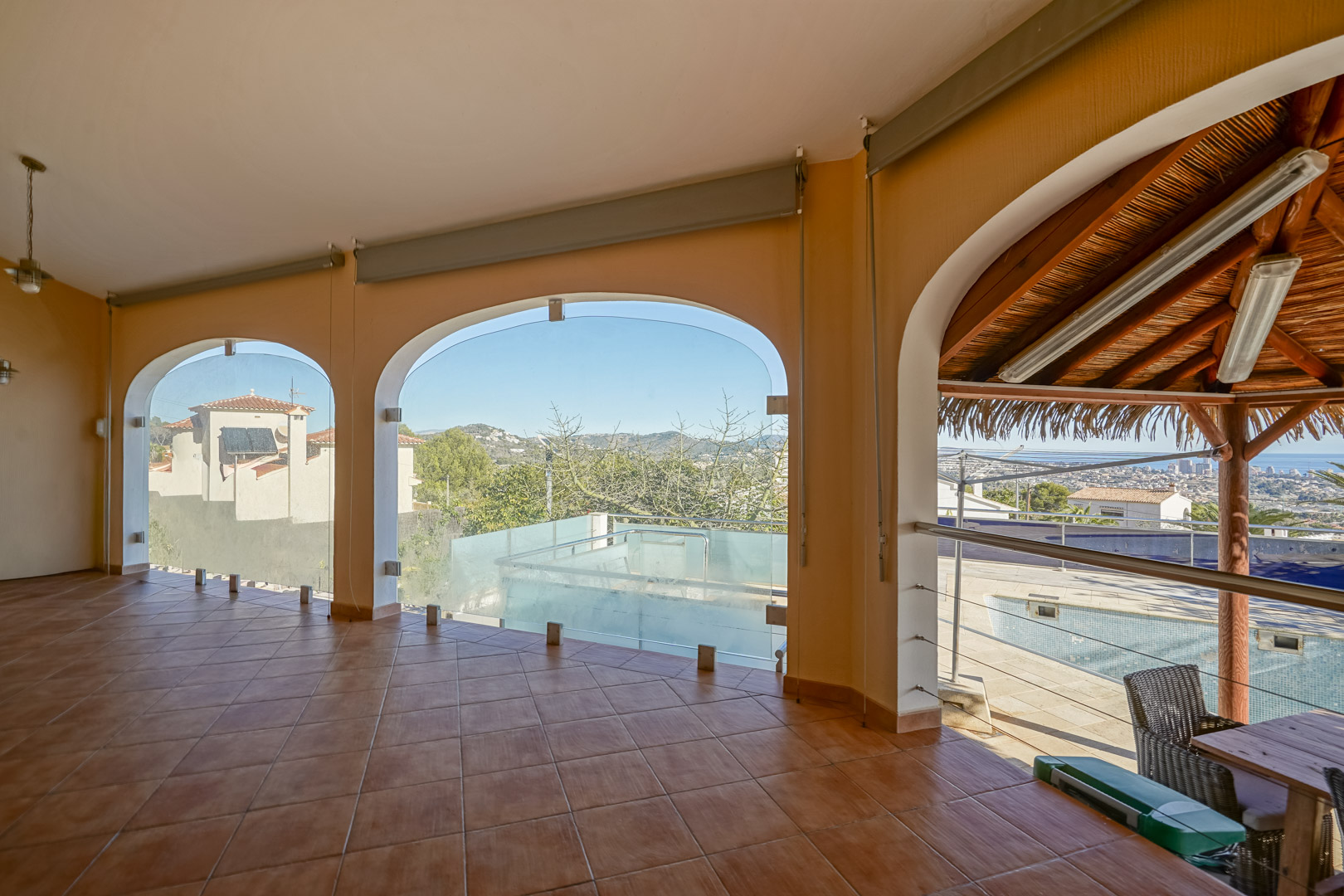 Überdachte Terrasse mit Terrakottaboden und verglasten Rundbögen, die einen Blick auf den Poolbereich und die Küste von Calpe bieten.