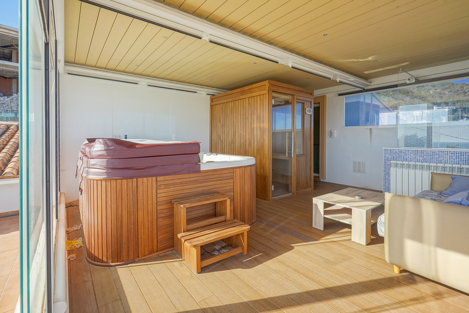 Verglaste obere Terrasse mit Wellnessbereich, ausgestattet mit einem holzverkleideten Whirlpool, einer Sauna und Holzboden unter einer Deckenkonstruktion mit Einbaustrahlern.