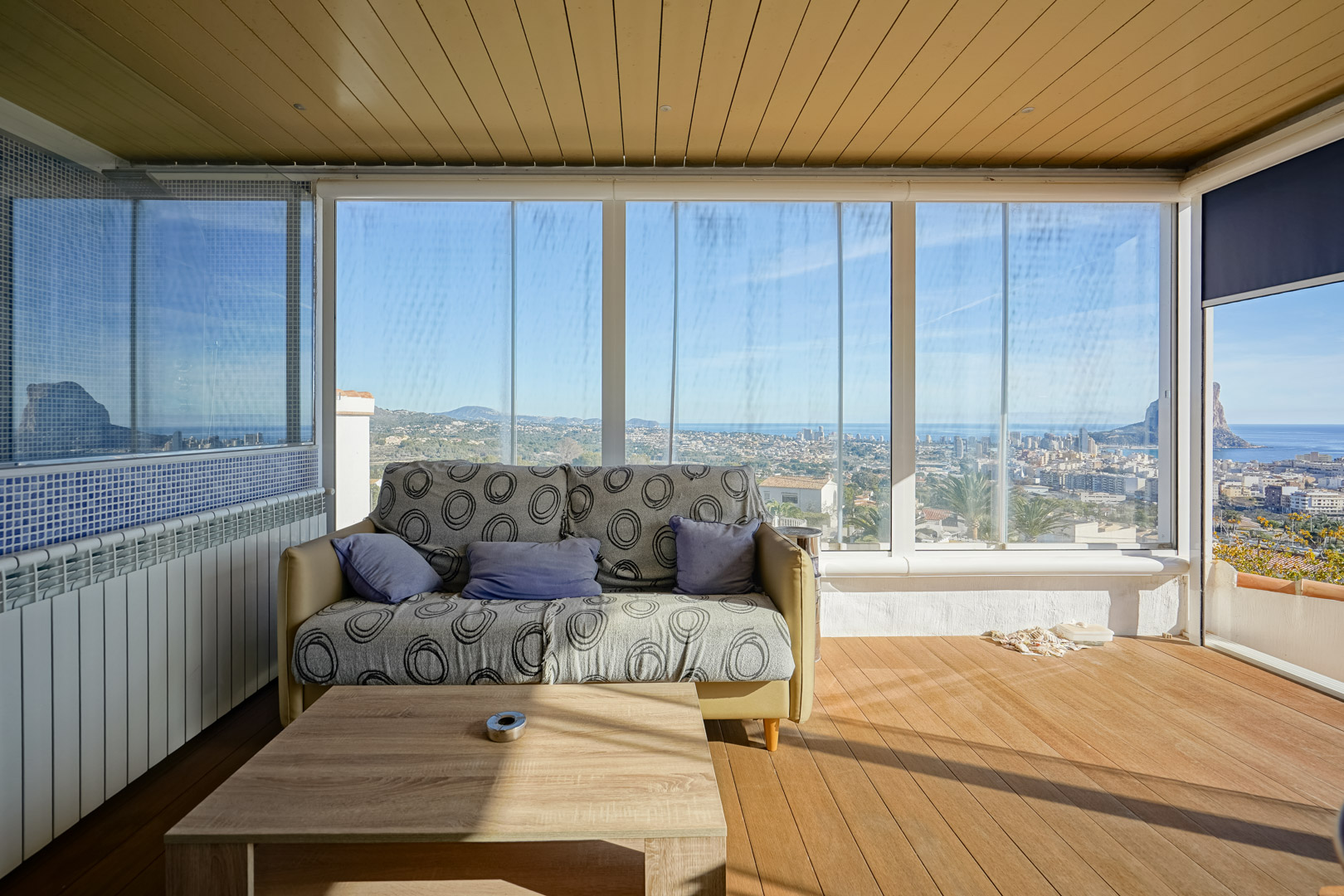 Verglaste Terrasse mit Glasschiebewänden, Holzboden und Panoramablick auf die Skyline von Calpe und den Peñón de Ifach. Mit holzgetäfelter Decke und Sitzbereich.
