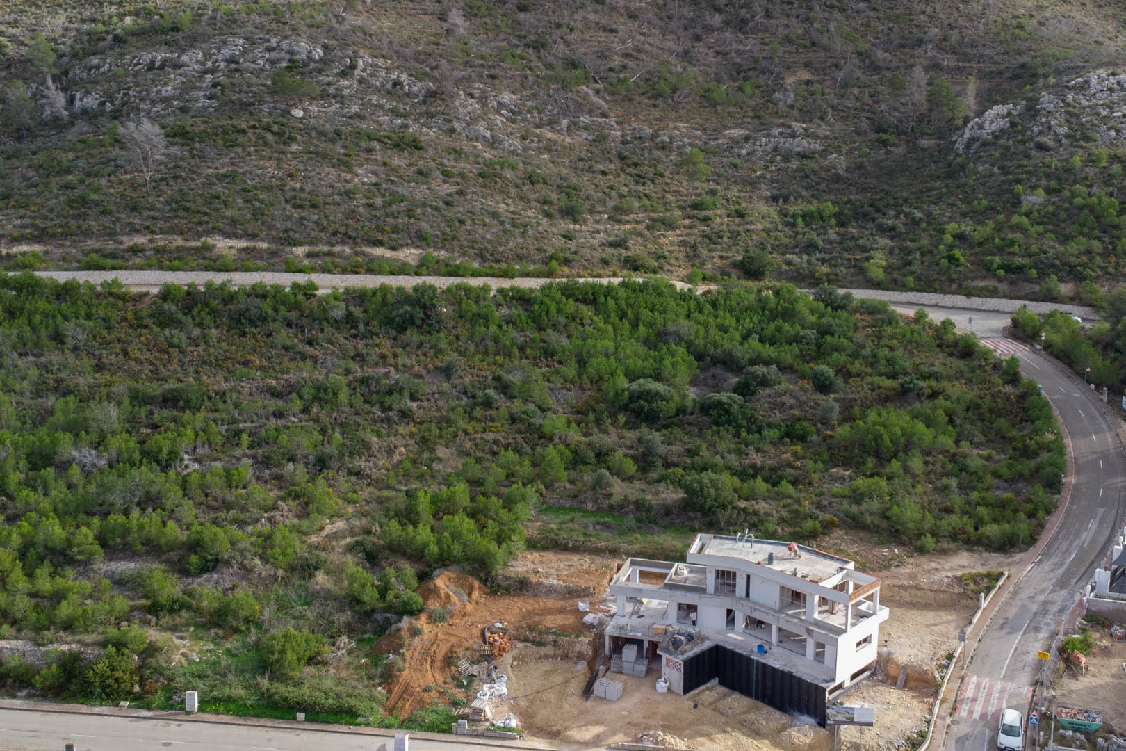 Luftaufnahme einer im Bau befindlichen Villa in Monte Olímpico, Jávea, umgeben von mediterraner Hanglage und asphaltierten Zufahrtsstraßen.