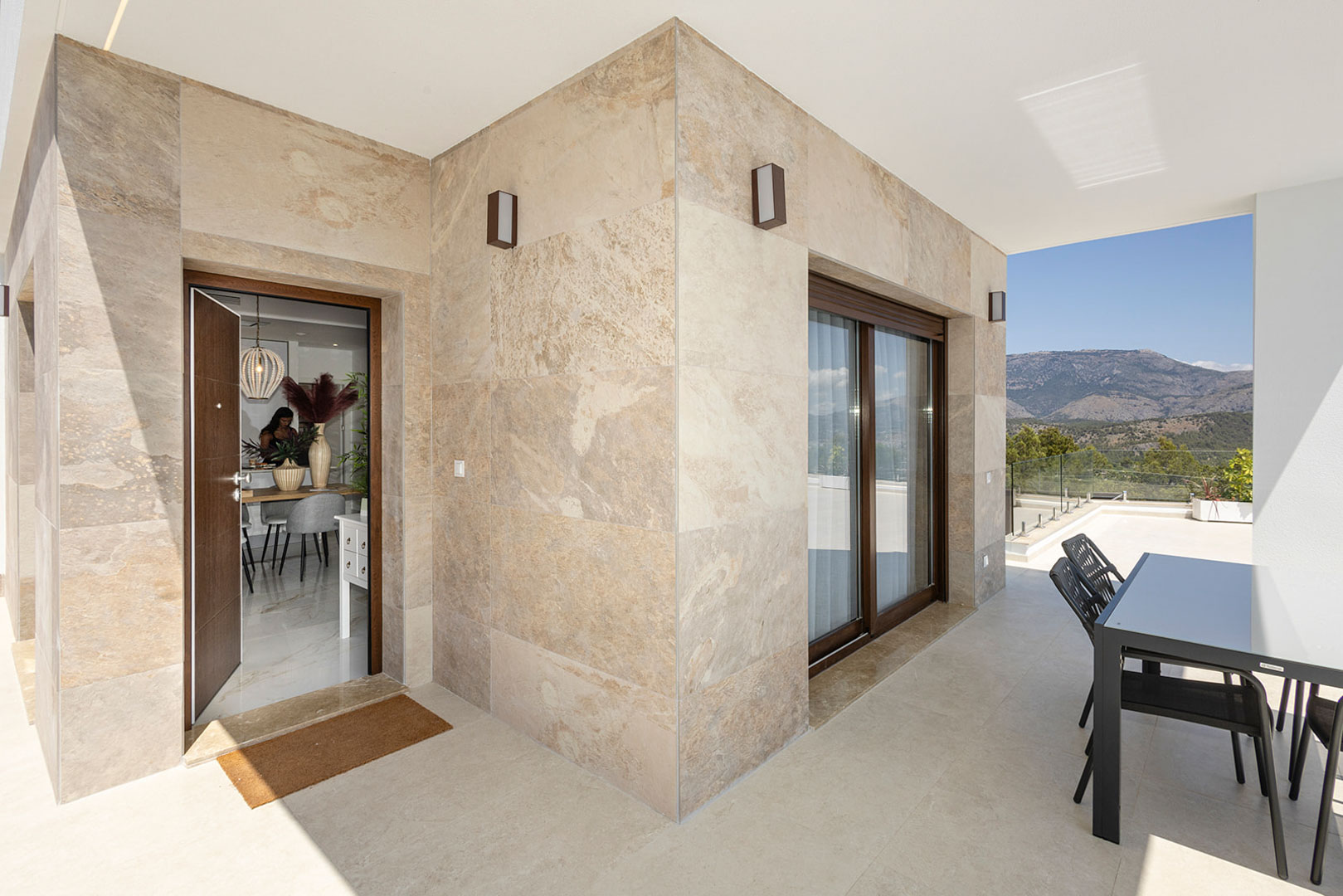 Außen-Terrasse mit Natursteinverkleidung, Essbereich und Panoramablick auf die Berge. Der Eingang führt direkt in den hellen Innenbereich der Villa.