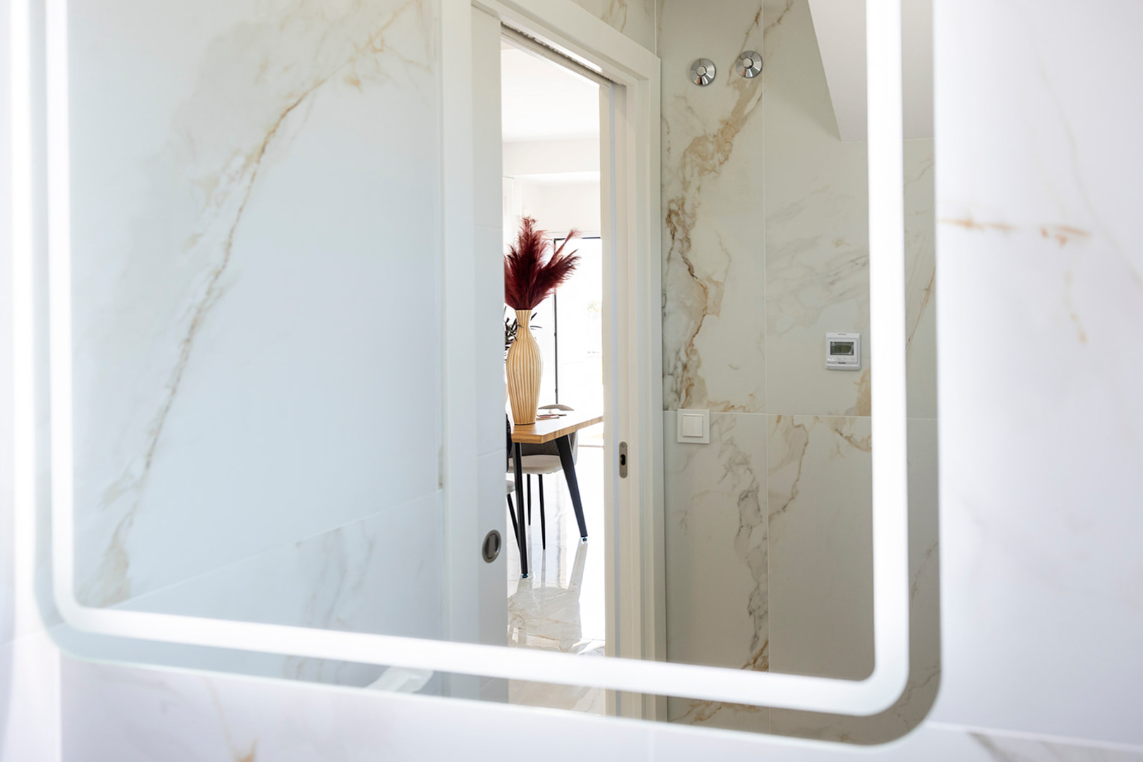 Badezimmer mit Wandfliesen in Marmoroptik und LED-hinterleuchtetem Spiegel, mit Blick durch die Tür auf den angrenzenden Essbereich.