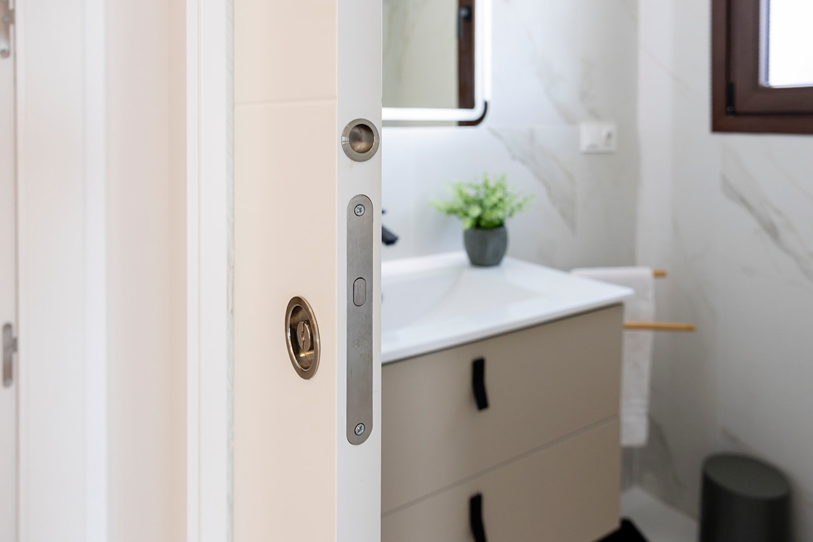 Detailansicht einer weißen Schiebetür mit bündigen Metallbeschlägen, die in ein modernes Badezimmer mit Wandfliesen in Marmoroptik und Waschtisch führt.