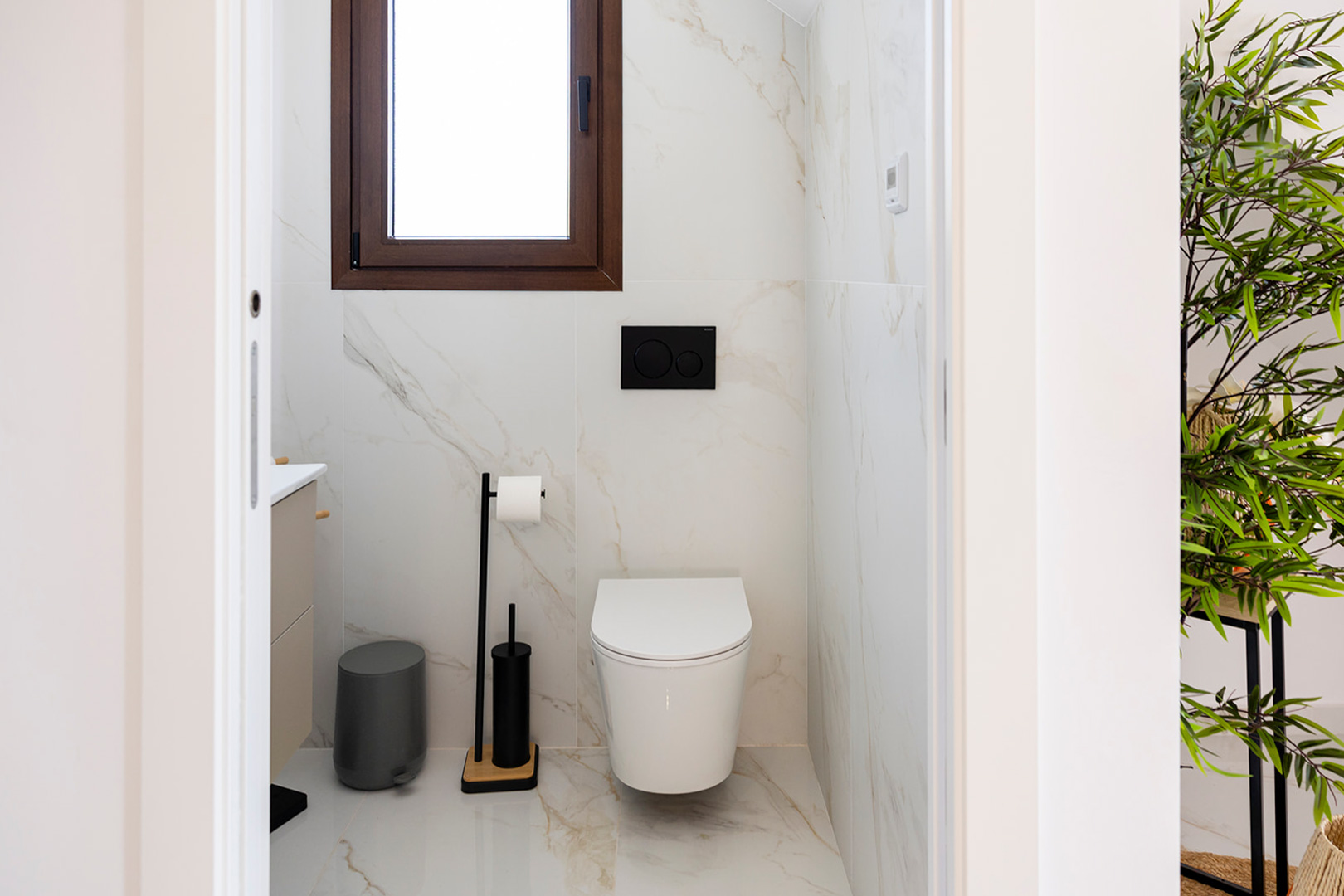 Gäste-WC mit großformatigen Fliesen in Marmoroptik, wandhängendem WC und schwarzen Akzenten. Ein Fenster mit Holzrahmen bietet Tageslicht.