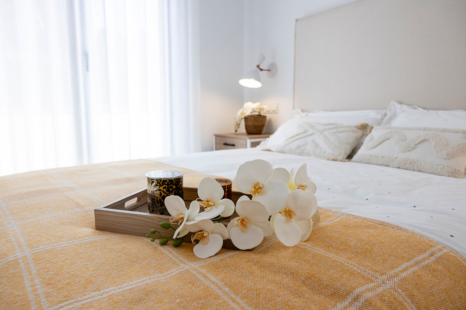Helles Schlafzimmer mit gepolstertem Kopfteil, Textilien in Weiß- und Senftönen sowie einem Dekotablett mit Orchideen. Natürliches Licht fällt durch die großen Fenster mit transparenten Vorhängen.
