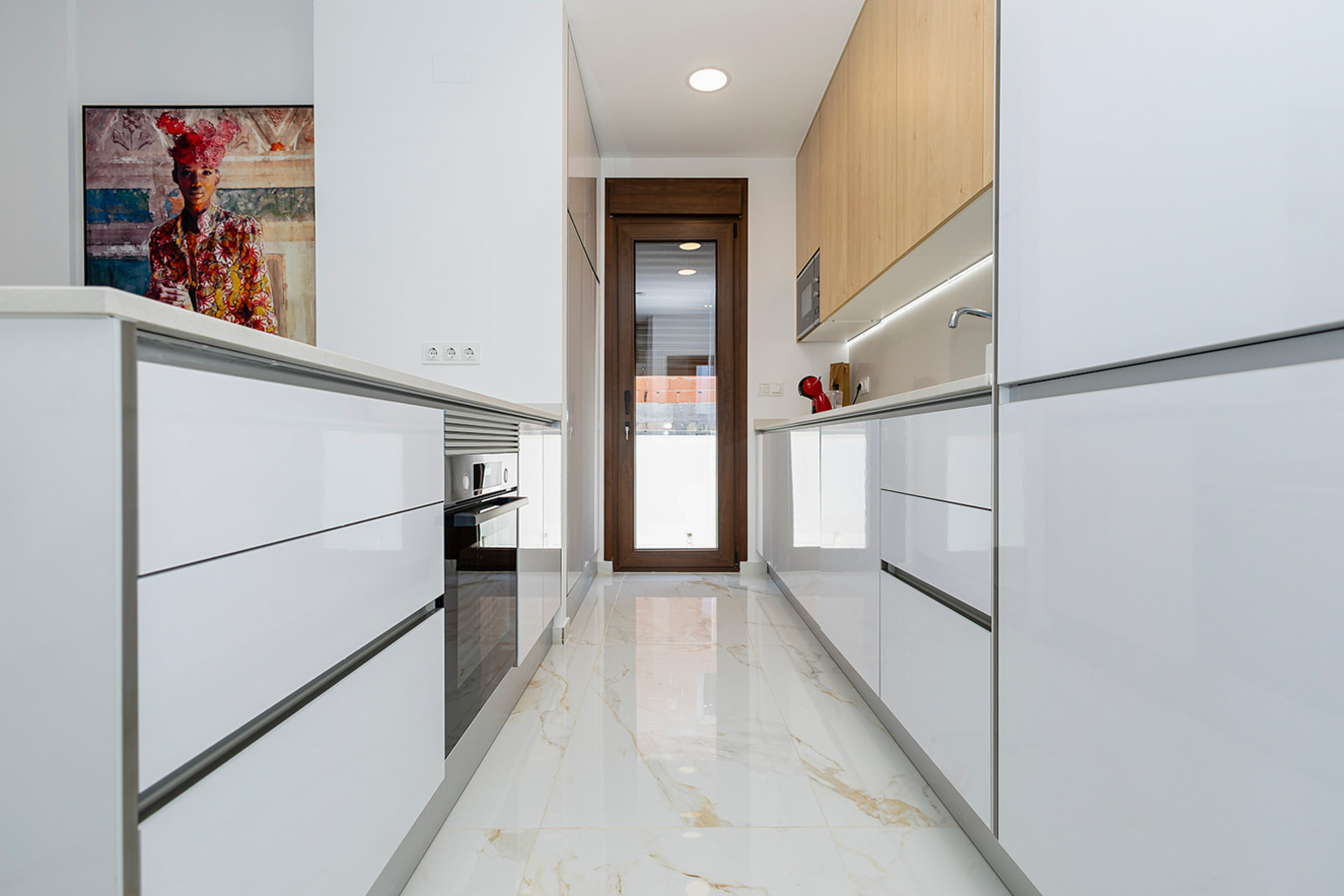 Moderne Küche mit hochglänzenden weißen Schränken, Oberteilen in Holzoptik und Feinsteinzeugboden in Marmoroptik. Ausgestattet mit Einbaugeräten und direktem Außenzugang.