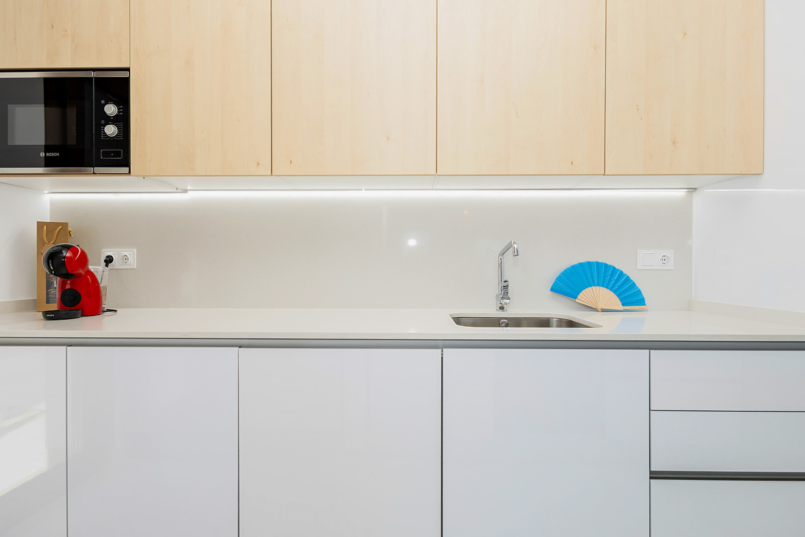 Moderne Küche mit Oberschränken in heller Holzoptik, weißen Unterschränken, Quarz-Arbeitsplatte und integrierter LED-Unterbaubeleuchtung.
