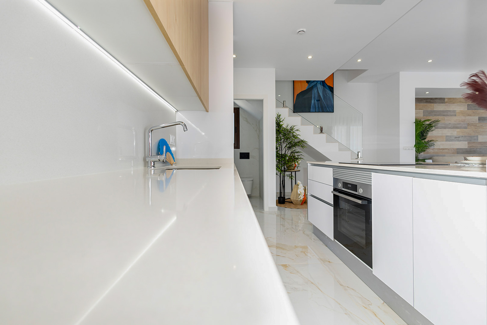 Moderne Küche mit weißer Arbeitsplatte, LED-Unterbaubeleuchtung und Kochinsel mit Backofen. Im Hintergrund ist die Treppe mit elegantem Glasgeländer sichtbar.