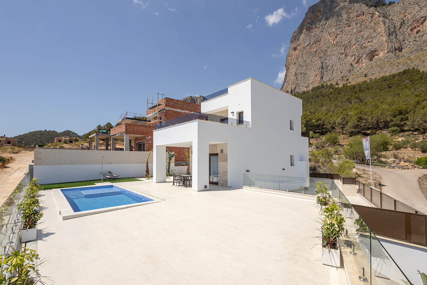 Moderne weiße Villa mit rechteckigem Pool, großzügiger Fliesenterrasse und Glasgeländern vor einer markanten Felskulisse.