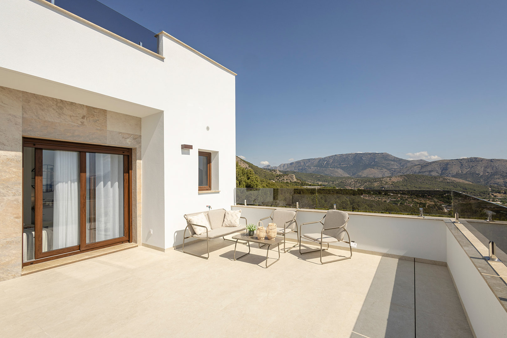 Obere Terrasse mit Glasgeländer, Gartenmöbeln und Panoramablick auf die Berge. Weiße Fassade mit Steinakzenten und hellem Keramikboden.