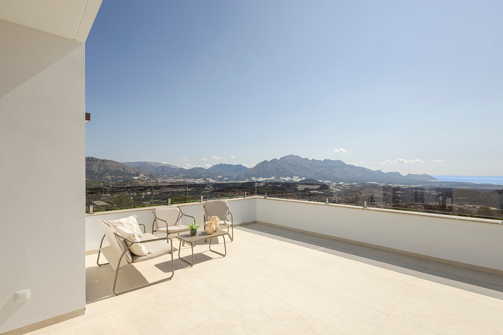 Obere Terrasse mit Glasgeländer, modernen Außenmöbeln und Panoramablick auf die Berge und das Meer. Ausgestattet mit großformatigen, hellen Bodenfliesen.