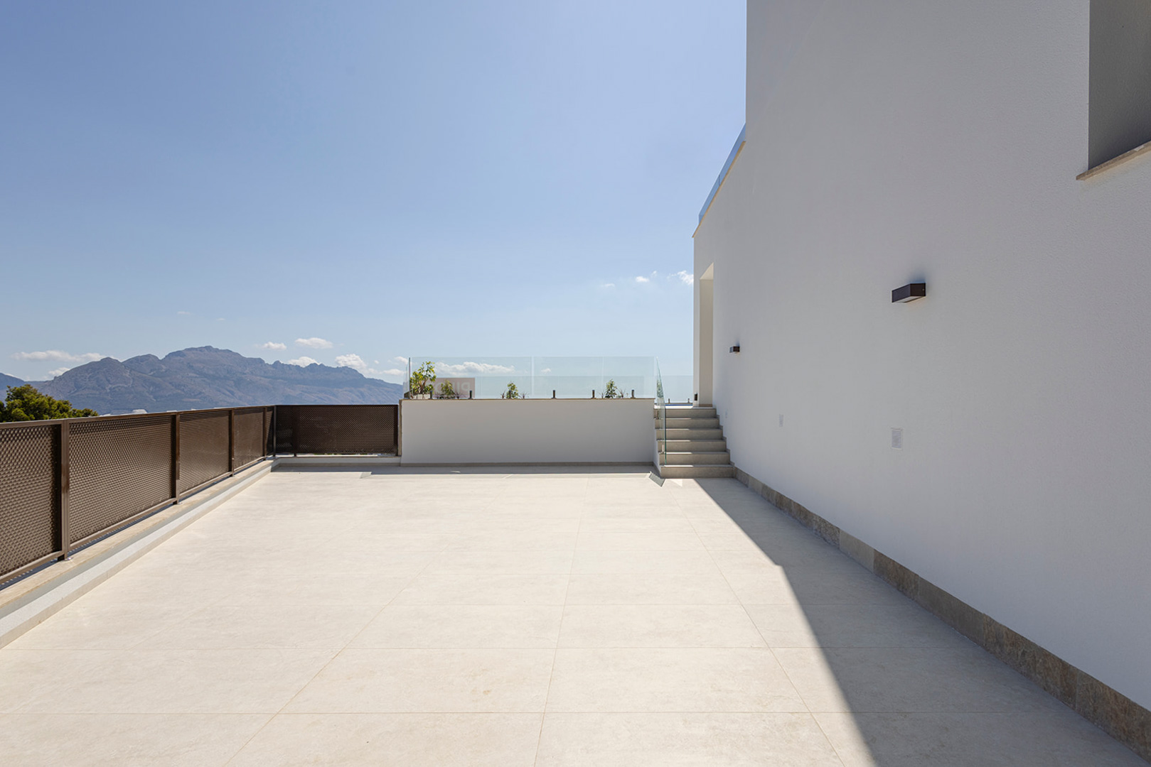 Obere Terrasse mit hellen Keramikfliesen, Glasgeländern und freiem Bergblick. Moderne Außenbeleuchtung an der minimalistischen weißen Fassade.