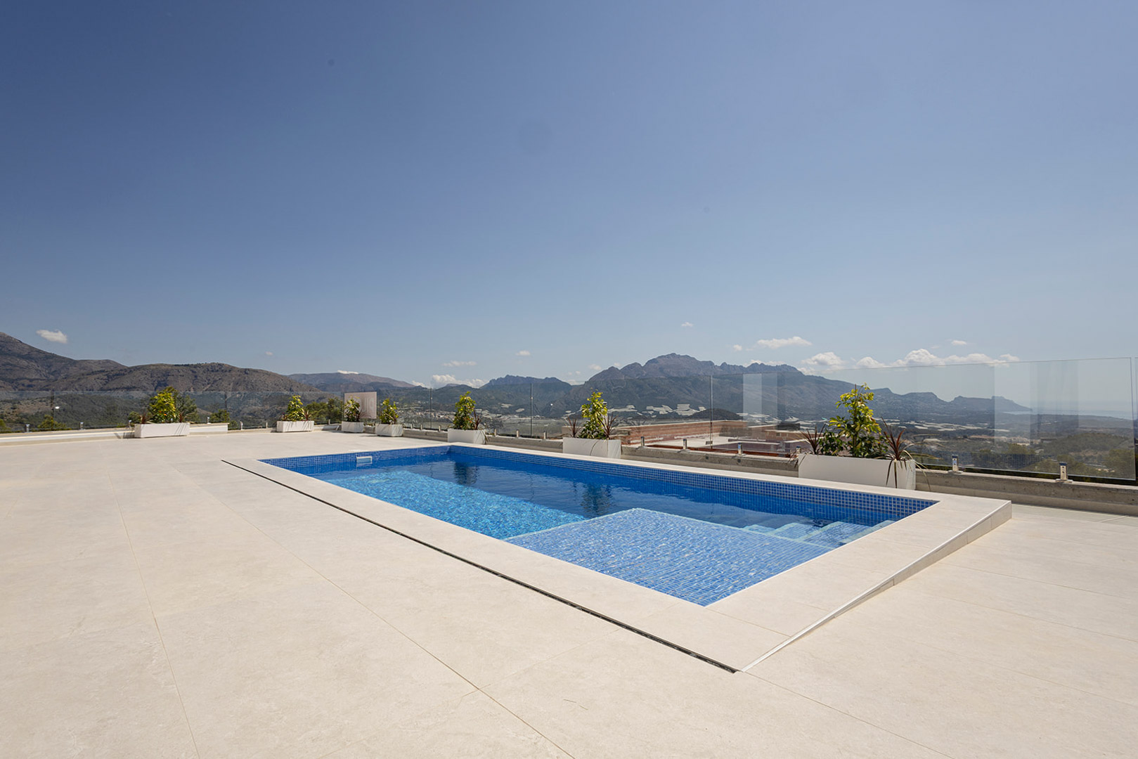 Privater Pool mit blauem Mosaik auf einer großzügigen hellen Terrasse, eingefasst von Glasgeländern mit Panoramablick auf die Berge und das Meer.