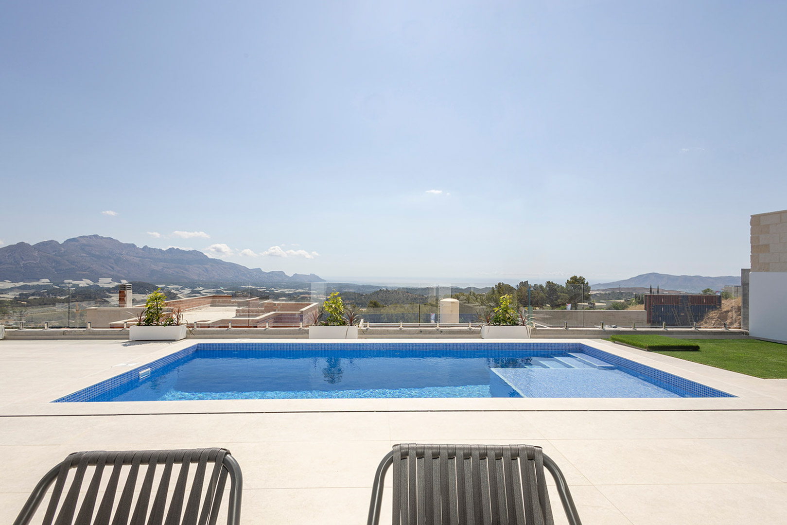 Privater Pool mit blauen Mosaikfliesen und integrierter Treppe, umgeben von einer hellen Steinterrasse und Glasgeländer mit Panoramablick auf Meer und Berge.