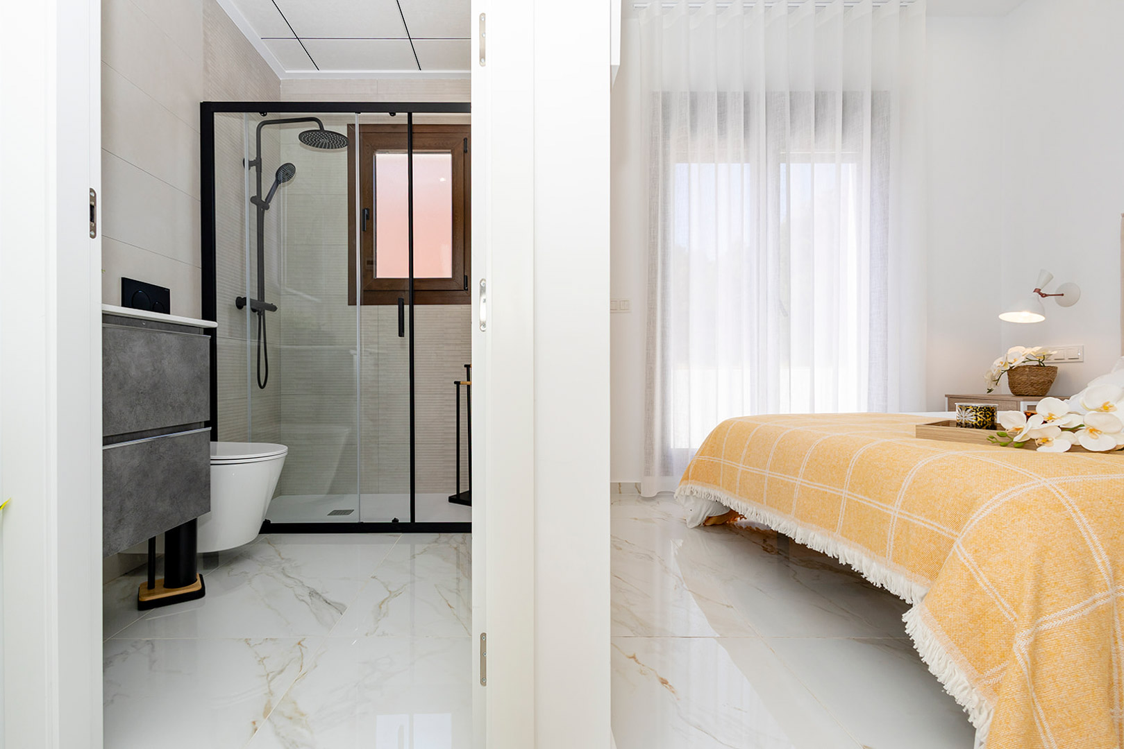 Schlafzimmer mit integriertem En-Suite-Bad, Feinsteinzeugböden in Marmoroptik und einer Dusche mit schwarzen Profilen und Glaswand.