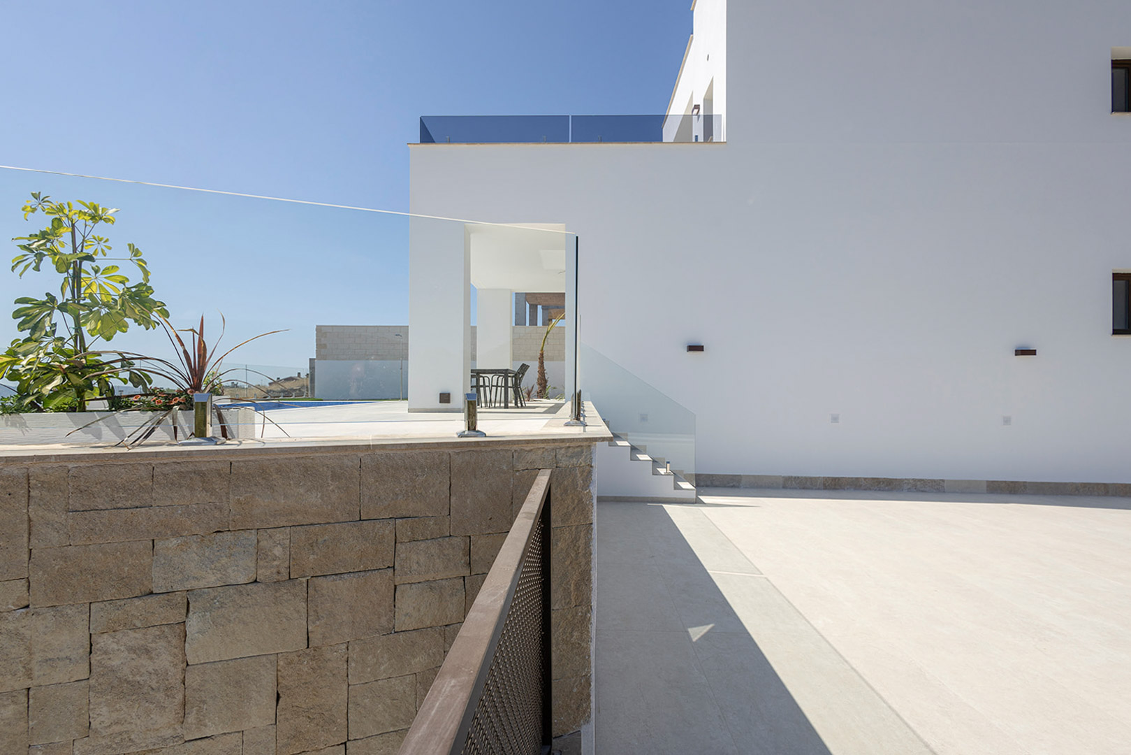 Weiße moderne Fassade mit Natursteinmauer, Glasgeländern und gepflastertem Außenbereich, der zur oberen Terrassenebene führt.