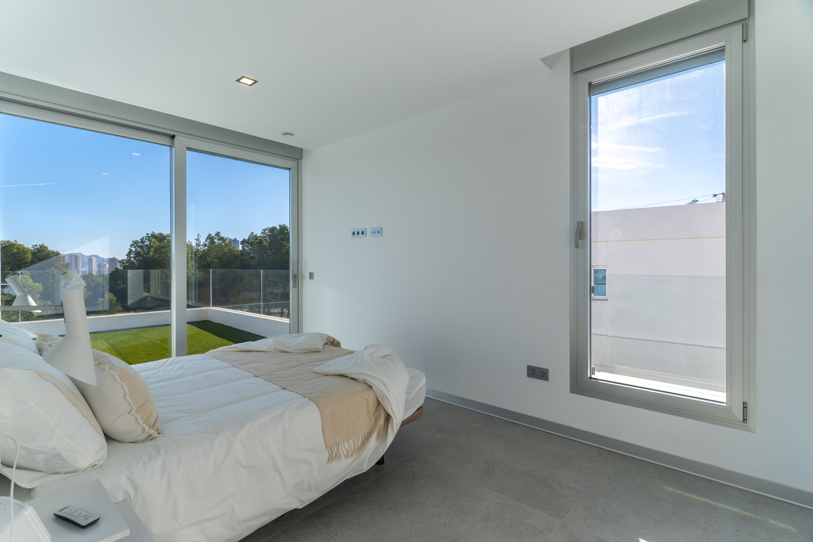 Helles Schlafzimmer mit grauen Fliesenböden, großen Aluminium-Schiebefenstern und direktem Zugang zu einer privaten Terrasse mit Kunstrasen und Weitblick.
