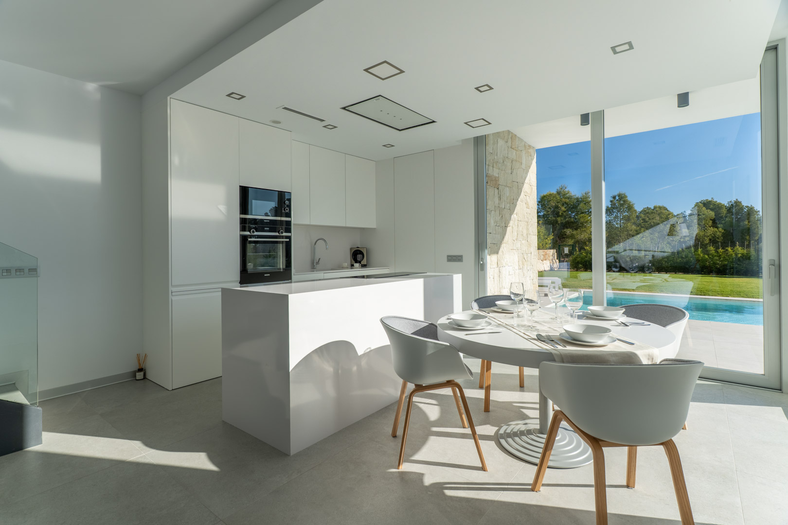 Minimalistische weiße offene Küche mit Kochinsel und Essbereich, ergänzt durch bodentiefe Fenster mit Blick auf den privaten Pool und Garten.