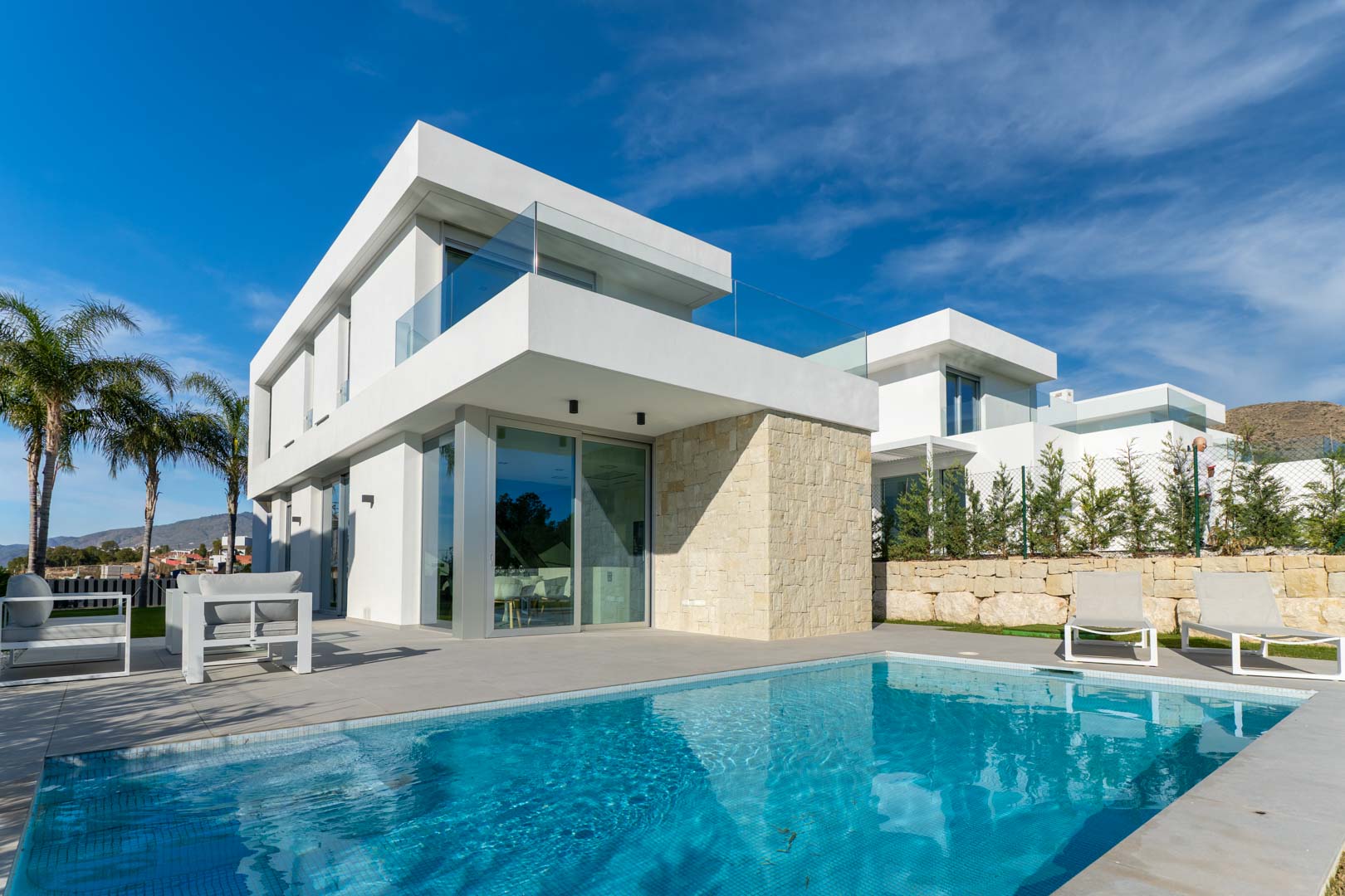 Moderne Villa mit privatem Pool, minimalistischer weißer Fassade, Natursteinwand und Terrassen mit Glasgeländern in sonniger Lage.
