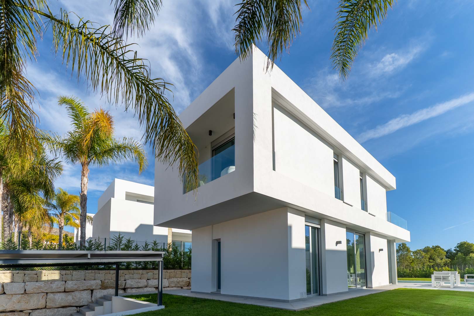 Moderne zweistöckige Villa mit auskragendem Obergeschoss, minimalistischer weißer Fassade und Glasgeländern. Der Außenbereich bietet Rasenflächen und Palmen.