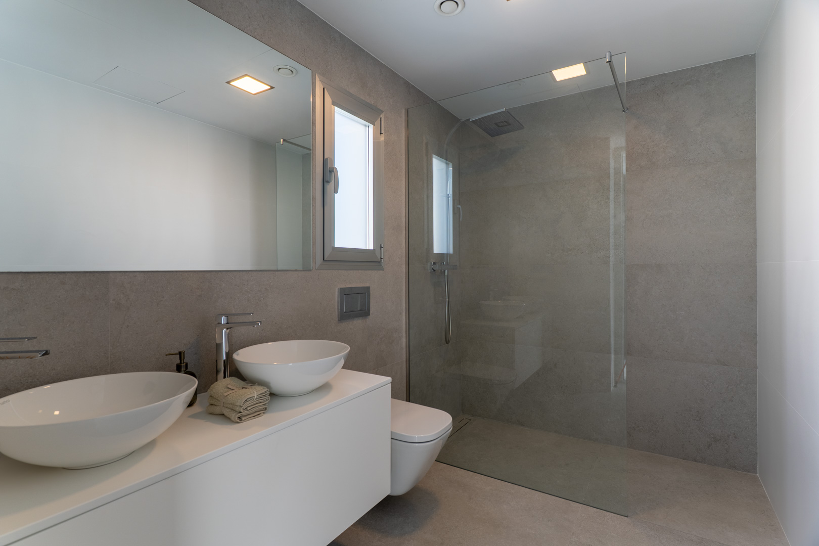 Modernes Badezimmer mit grauen Feinsteinzeugfliesen, Doppelwaschbecken auf weißem Unterschrank und begehbarer Dusche mit rahmenloser Glaswand.