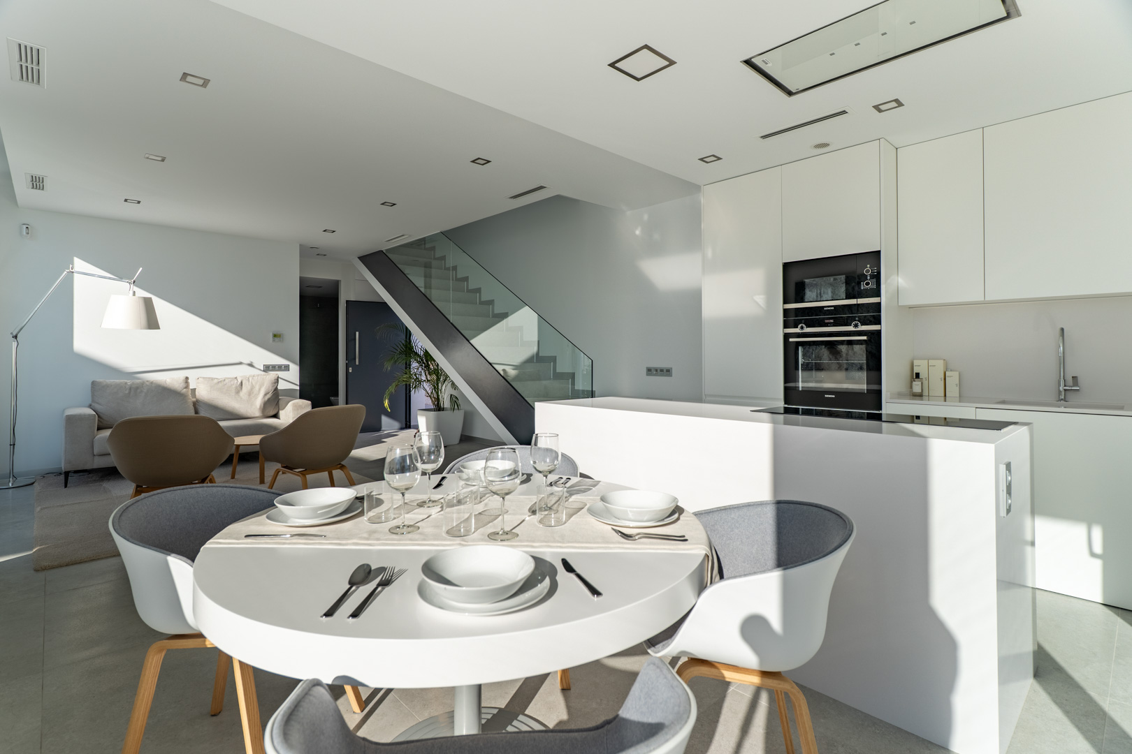 Offener Wohnbereich mit minimalistischer weißer Küche, Kochinsel und integrierten Geräten. Eine moderne Treppe mit Glasgeländer prägt das architektonische Design.