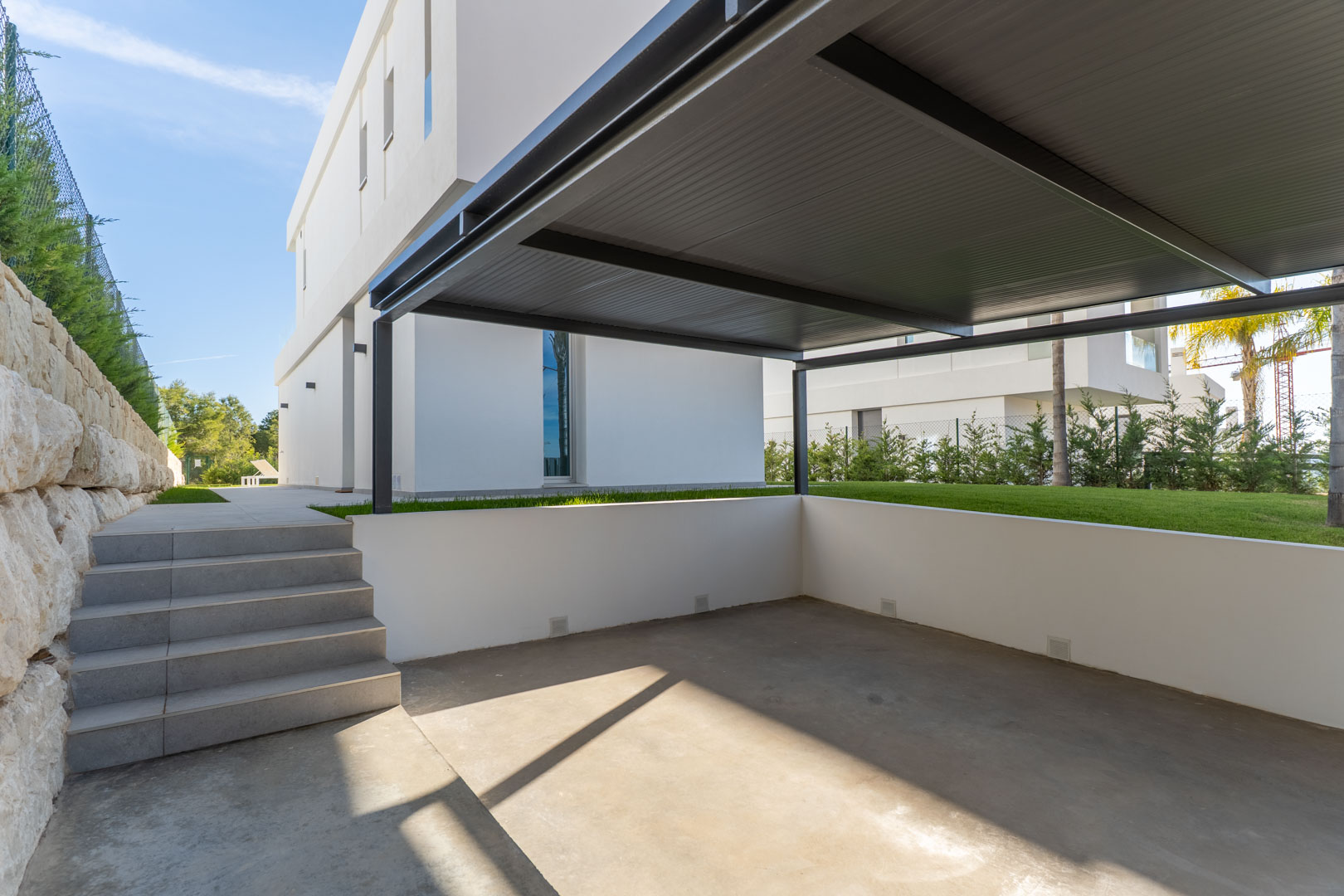 Privater Parkplatz mit Metall-Carport, Betonboden und gefliesten Stufen, die zum Gartenbereich und zum modernen Villengebäude führen.