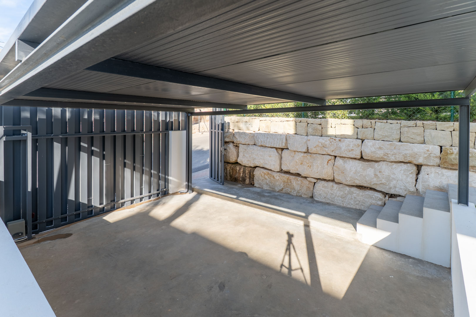 Überdachter Privatparkplatz mit Metallkonstruktion, Betonboden und einer Natursteinmauer sowie integrierten Stufen zum Wohnbereich.