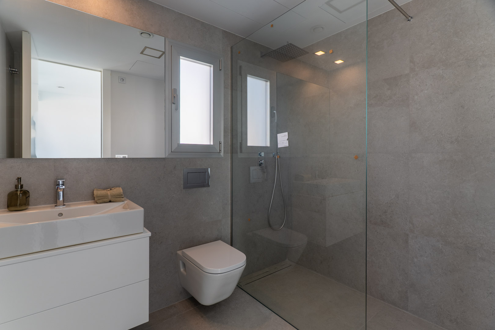 Zeitgenössisches Badezimmer mit grauen Fliesen, begehbarer Dusche mit Glaswand, wandhängendem WC und integriertem Waschbecken auf weißem Unterschrank.