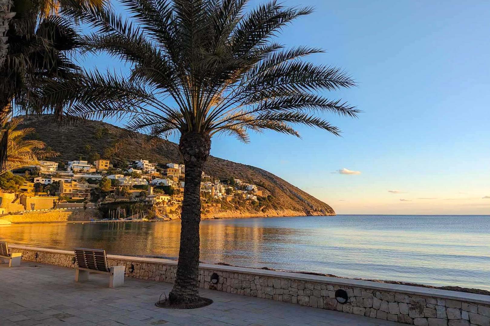 Blick auf die Promenade von El Portet in Moraira mit Palmen, Natursteinmauern und weißen Villen, die sich an den Küstenhang schmiegen.